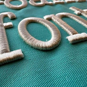 May include: Close-up of embroidered letters spelling "FONT" in a light beige thread on a teal fabric background. The letters are thick and have a textured appearance, with the thread creating a raised effect. The fabric has a diagonal weave.