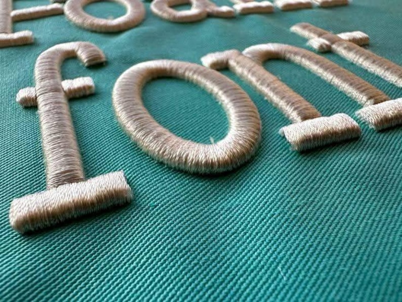 May include: Close-up of embroidered letters spelling "FONT" in a light beige thread on a teal fabric background. The letters are thick and have a textured appearance, with the thread creating a raised effect. The fabric has a diagonal weave.