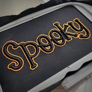 May include: Black fabric with the word "Spooky" embroidered in black with a yellow outline. The embroidery is in a bold, stylized font. The fabric is stretched within an embroidery hoop.