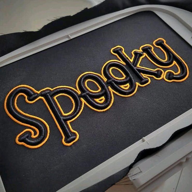 May include: Black fabric with the word "Spooky" embroidered in black with a yellow outline. The embroidery is in a bold, stylized font. The fabric is stretched within an embroidery hoop.