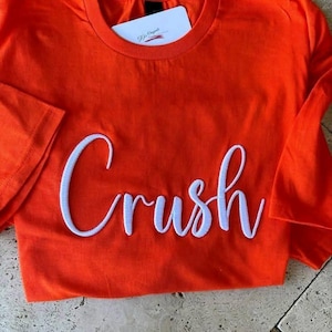 May include: Bright orange t-shirt with the word "Crush" embroidered in white cursive script across the front. A white tag is tucked into the neckline. The shirt is folded, showing the sleeve.