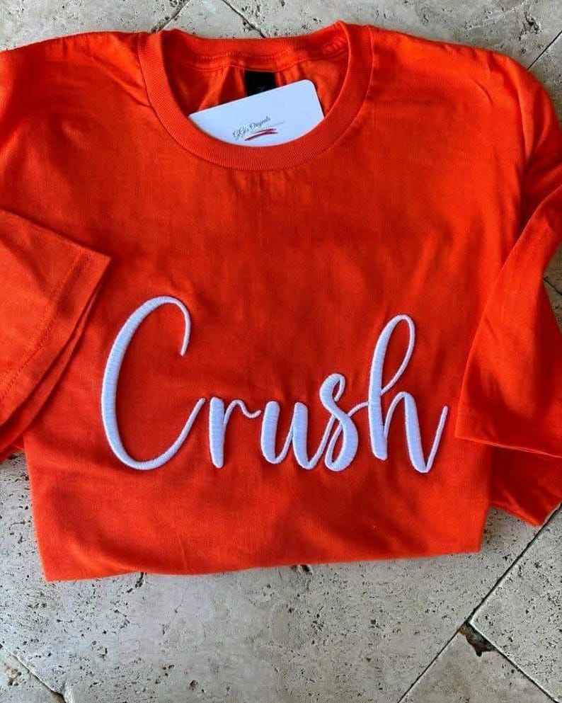 May include: Bright orange t-shirt with the word "Crush" embroidered in white cursive script across the front. A white tag is tucked into the neckline. The shirt is folded, showing the sleeve.