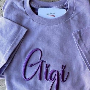 May include: A lavender-colored t-shirt with the name "Gigi" embroidered in a darker purple script across the front. The shirt has short sleeves and a crew neck. A white card with text is tucked into the neckline.