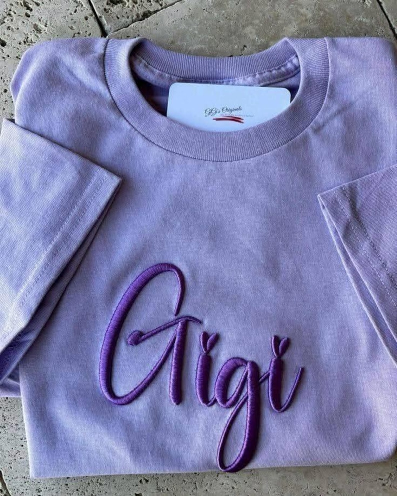 May include: A lavender-colored t-shirt with the name "Gigi" embroidered in a darker purple script across the front. The shirt has short sleeves and a crew neck. A white card with text is tucked into the neckline.