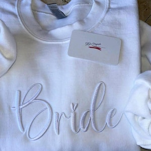 May include: White crewneck sweatshirt with the word "Bride" embroidered in a cursive font. A small tag with the text "Gg's Originals" is placed on the sweatshirt. The sweatshirt is folded.