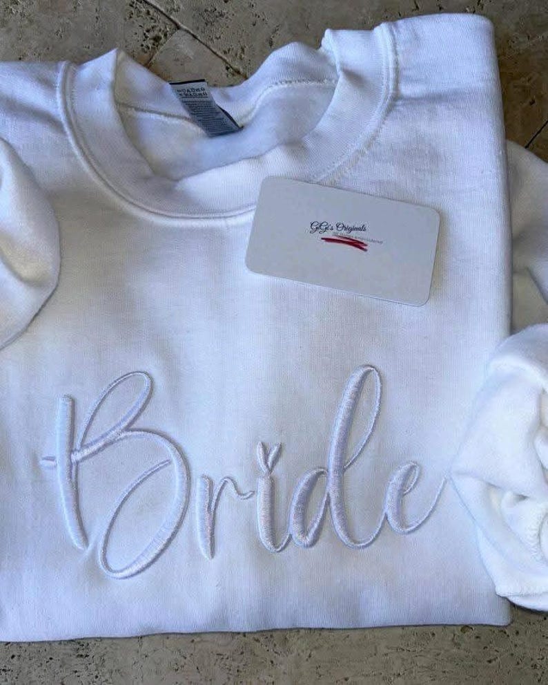 May include: White crewneck sweatshirt with the word "Bride" embroidered in a cursive font. A small tag with the text "Gg's Originals" is placed on the sweatshirt. The sweatshirt is folded.