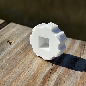 May include: A white, gear-shaped object with a square center hole and ridged edges. The object is made of a solid material and sits on a weathered wooden surface, casting a shadow. The design suggests a mechanical or industrial component.