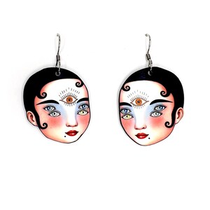 May include: A pair of unique earrings featuring a stylized face design. Each earring has a black background with a face illustration. The face has multiple eyes, red lips, and a beauty mark. The earrings are suspended from silver-colored hooks.