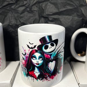 May include: White ceramic mug featuring a colorful illustration of Jack Skellington and Sally from The Nightmare Before Christmas. The design includes bats and splashes of pink, blue, and purple. The mug is sitting on a white box.
