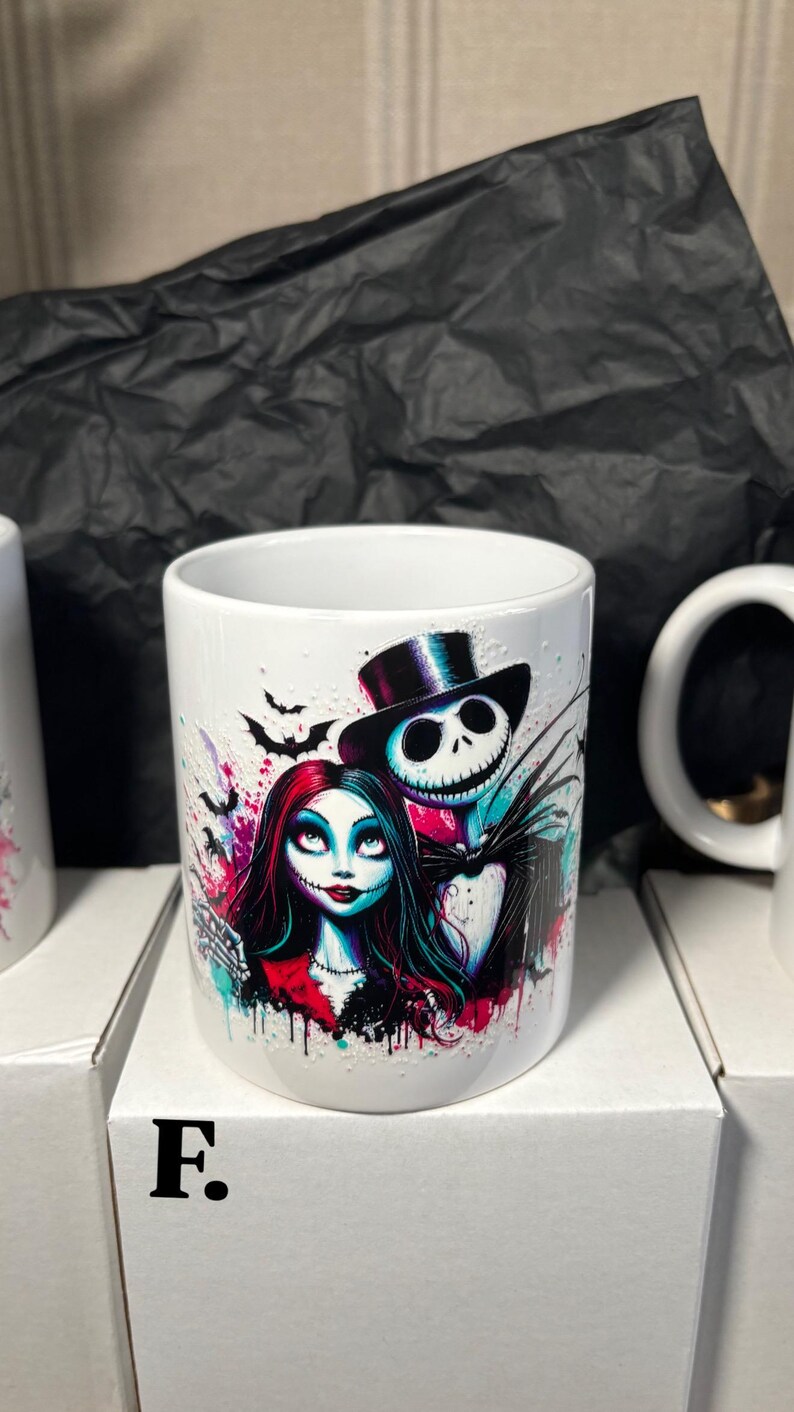 May include: White ceramic mug featuring a colorful illustration of Jack Skellington and Sally from The Nightmare Before Christmas. The design includes bats and splashes of pink, blue, and purple. The mug is sitting on a white box.
