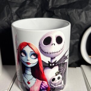 May include: White ceramic mug featuring Jack Skellington and Sally from The Nightmare Before Christmas. Sally has red hair and a stitched appearance. Jack has a skeletal face. A small pumpkin is also featured on the mug.