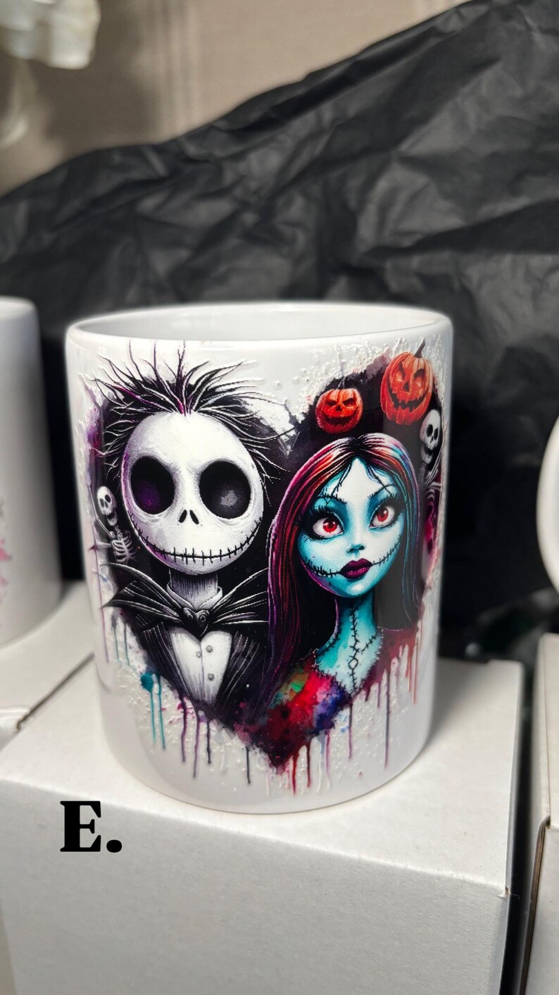 May include: White ceramic mug featuring a heart-shaped design with Jack Skellington and Sally from The Nightmare Before Christmas. The design includes pumpkins, skulls, and a watercolor effect. The letter "E." is visible on the mug.