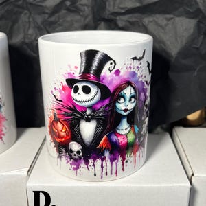 May include: White ceramic mug featuring Jack Skellington and Sally from The Nightmare Before Christmas, with a pumpkin and skull. The design includes a black top hat, bats, and colorful paint splatters. A Halloween-themed item.