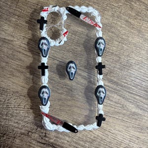 Op de afbeelding: Transparante telefoonhoes versierd met witte frosting-achtige rand, zwarte kruisen en kleine Scream-maskerbedels. Miniatuur bloederige messen zijn ook bevestigd. De hoes is ontworpen voor een telefoon.
