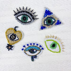 Peut inclure: Cinq broches en forme d'œil, ornées de perles, aux designs variés. L'une est ornée de pointes, une autre est triangulaire. Les couleurs incluent le vert, le bleu, l'or et l'argent. Les broches sont décorées de perles et de paillettes.