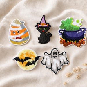 May include: Five embroidered Halloween patches featuring a candy corn character, a black cat wearing a purple witch hat, a purple cauldron with green bubbling liquid over a fire, a yellow full moon with black bats, and a white ghost.