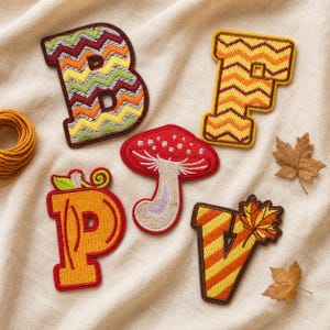 Peut inclure: Un ensemble de cinq écussons brodés représentant les lettres B, P et V, un champignon et la lettre F. Les écussons présentent des couleurs automnales incluant orange, jaune, rouge et marron, avec des motifs en chevron, une tige de citrouille.