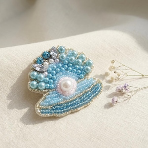 May include: A handcrafted, beaded seashell brooch in shades of blue, white, and silver. The brooch features a large pearl at the center, surrounded by smaller pearls and crystals. The edges are outlined with silver beads, creating a detailed and elegant accessory.