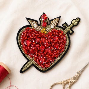 May include: A heart-shaped patch, heavily embellished with red beads and gemstones, pierced by a gold sword. The patch features a black border and a gold crown. A spool of red thread and gold scissors are in the background.
