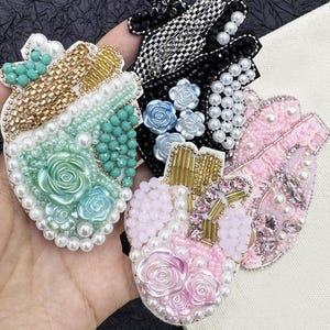 May include: A collection of heart-shaped brooches, each intricately embellished with beads, pearls, and sequins. The brooches feature various color schemes, including mint green, pink, and black, with floral and geometric patterns. The brooches are handmade.