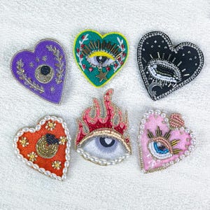 May include: Six heart-shaped embroidered patches in various colors. Designs include an eye, flames, and celestial elements. Embellishments include beads, pearls, and metallic thread. The patches are on a white background.