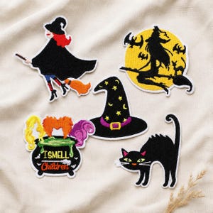 May include: Five embroidered patches featuring Halloween themes: a witch on a broom, a witch silhouette against a yellow moon, a black witch hat with yellow stars, a black cat with arched back, and a cauldron with the text I SMELL CHILDREN.