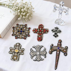 May include: An assortment of decorative crosses in various designs and sizes. Some crosses are embellished with colorful gemstones, while others are made of clear crystal. A white book and flowers are in the background.