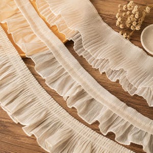 May include: A length of off-white, semi-sheer pleated fabric trim featuring a ruffled edge. The material has a fine, delicate texture with consistent vertical pleats running along the length of the ribbon, creating a soft, wavy, and layered appearance.