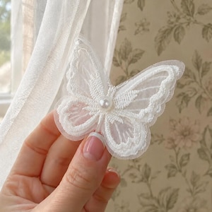 May include: A white butterfly applique crafted from sheer mesh fabric with intricate embroidered edges and a single central pearl bead accent. The design features delicate stitching patterns that define the wings, creating a layered appearance.