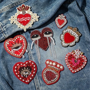 May include: A collection of heart-shaped embroidered patches with intricate beadwork and embellishments. The patches feature red, white, and gold colors, with designs including eyes, crowns, and tear-like accents. They are displayed on a denim jacket.