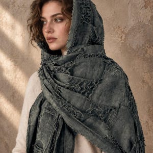 May include: A dark gray, distressed-look scarf with a frayed, open-weave texture draped over the head and shoulders. The fabric features a patchwork-style construction with visible seams and raw edges, worn over a light-colored, long-sleeved sweater.