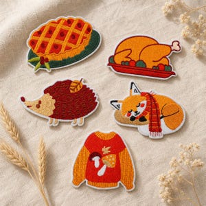 May include: Five embroidered patches featuring a lattice-top pie, a roasted turkey on a platter, a brown hedgehog with a leaf, a sleeping fox wearing a red scarf, and a red sweater with mushroom designs, all arranged on a textured fabric surface.