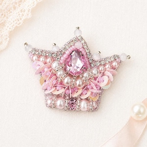 May include: A handcrafted, pink and silver crown brooch. The crown is adorned with a large pink gemstone, pearls, sequins, and small crystals. The brooch is set against a white background with a lace trim.