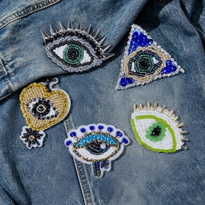 May include: A collection of beaded eye-shaped patches in various designs. The patches feature intricate beadwork in colors like blue, green, gold, and silver, with some incorporating spikes and a heart shape. They are displayed on a denim jacket.
