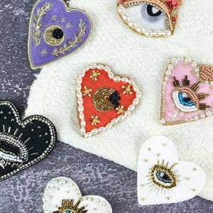 Peut inclure: Plusieurs patchs brodés en forme de cœur, de couleurs variées : violet, rouge, noir, rose et blanc. Chaque patch présente des broderies et des ornements complexes, avec des motifs d'yeux et floraux. Les patchs sont exposés sur une surface blanche texturée.