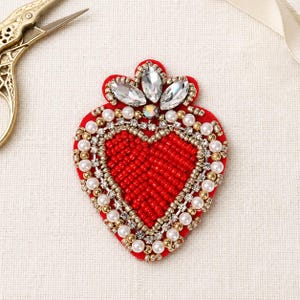 May include: A heart-shaped, red beaded applique with a crown of clear crystals. The applique is embellished with pearls and gold-colored beads. A pair of gold scissors is visible in the background.