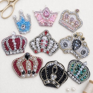 May include: A collection of nine decorative crown patches in various colors and designs. The crowns are embellished with beads, sequins, and rhinestones. Colors include blue, pink, red, black, and silver. Some crowns have pearl accents.
