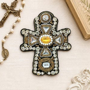May include: A black, beaded cross with a central yellow oval gem and clear, triangular, and round gems. The cross is surrounded by gold-colored beads. A rosary and a book are in the background.