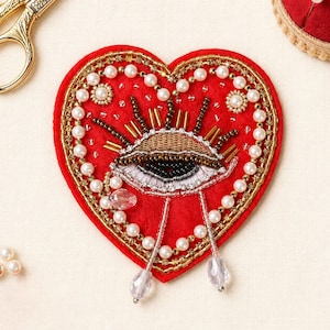 May include: A red heart-shaped patch with an eye design, adorned with pearls, gold beads, and clear crystal accents. The eye features detailed embroidery in black, brown, and white. Decorative craft item.