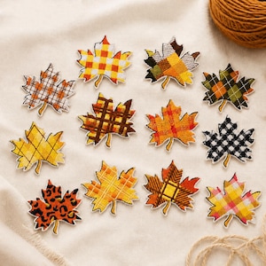 May include: A collection of twelve embroidered maple leaf patches featuring various patterns including yellow, orange, and brown plaid, black and white gingham, and an orange leopard print. Each leaf-shaped patch has a distinct stitched border.