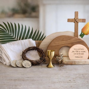 May include: A wooden cross with a rope detail, a yellow tulip, and a wooden display with the text "He is not here, for He is risen, just as He said, Matthew 28:6." Also, a gold chalice, a small rooster statue, a crown of thorns, and three silver coins.