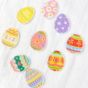 Peut inclure: Collection de neuf écussons en forme d'œufs de Pâques colorés. Les motifs comprennent des fleurs, des formes géométriques et l'inscription "HAPPY EASTER" sur un œuf jaune. Les œufs sont de couleurs jaune, rose, violet, vert et rouge.