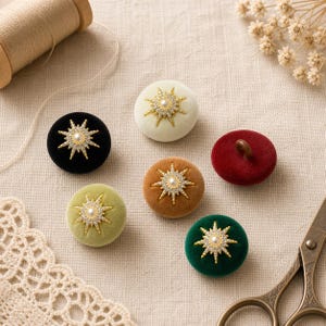 May include: A collection of six round velvet buttons in black, cream, light green, brown, dark red, and dark green. Five buttons feature a gold-tone starburst emblem with a central faux pearl and clear crystals. One red button is plain.