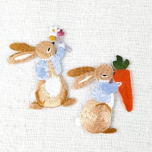 Peut inclure: Deux écussons brodés de lapins. L'un tient un bouquet de fleurs, l'autre une grosse carotte orange. Les deux lapins portent des vestes bleues et ont des oreilles marron et un corps beige. Les écussons sont sur fond blanc.