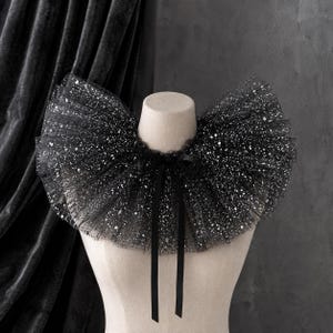 May include: A black ruffled collar made of sheer tulle fabric featuring a dense pattern of silver glitter throughout. The collar is gathered in a wide, circular shape around the neck and is secured at the front with a thin black satin ribbon tie.