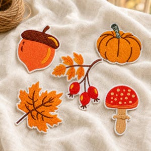 May include: A set of five embroidered patches featuring autumn themes: an orange acorn with a brown cap, a round orange pumpkin, a branch with yellow leaves and red berries, a jagged orange maple leaf, and a red mushroom with white spots.