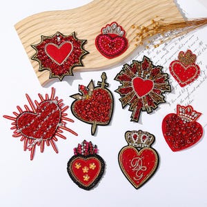 May include: A collection of red heart-shaped embroidered patches with various designs. Some hearts feature crowns, sunbursts, or decorative elements. The patches are embellished with beads and sequins, and outlined with gold or black thread.