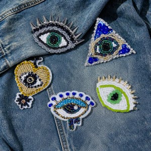 May include: A collection of beaded eye-shaped patches in various designs. The patches feature intricate beadwork in colors like silver, gold, blue, and green, with some incorporating spikes and a heart shape. They are displayed on a denim jacket.