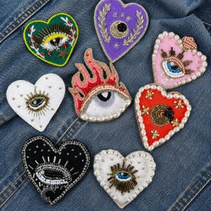 May include: A collection of heart-shaped embroidered patches with eye and flame designs. The patches feature various colors, including green, purple, pink, red, white, and black, and are embellished with beads and pearls. They are arranged on a blue denim background.