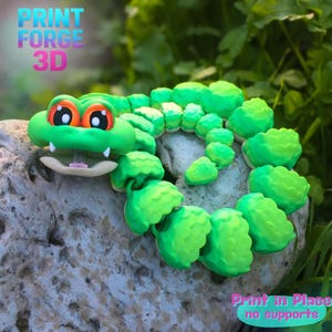 May include: A bright green 3D-printed articulated alligator toy with orange eyes and white teeth. The text "PRINT FORGE 3D" is in the upper left corner, and "Print in Place no supports" is in the lower right corner.